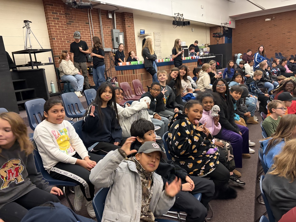 A large group of elementary students sit in rows of chairs inside an auditorium, smiling and waving toward the camera as they wait for a performance to begin. Behind them, older students and staff prepare near the back wall where stage lights, sound equipment, and colorful props are visible.