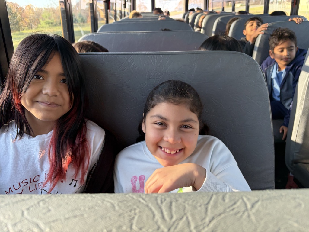 Two students sit side by side on a school bus, smiling warmly toward the camera. Sunlight filters through the windows behind them, and several classmates can be seen chatting in the background.