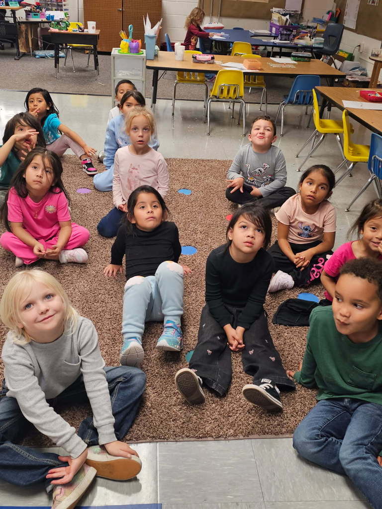 “Students sitting cross-legged on a brown rug, looking toward the camera in a kindergarten classroom with tables and chairs behind them.”