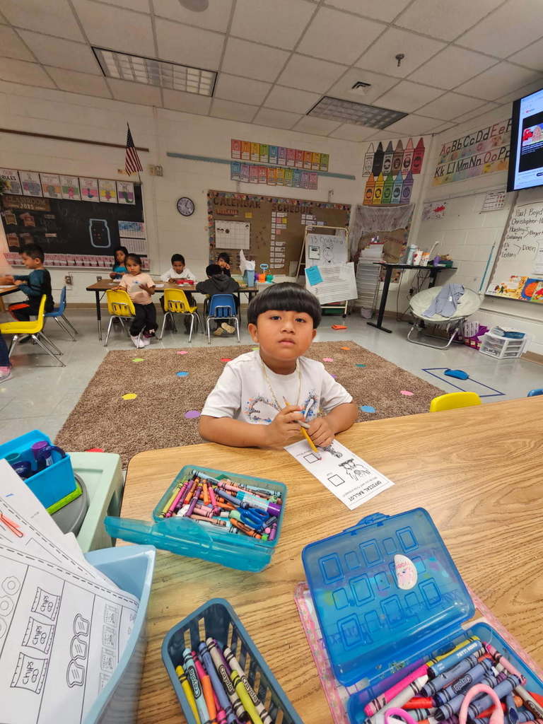 “Student at a front table working on a worksheet with a pencil; open crayon boxes on the table and classmates working in the background.”