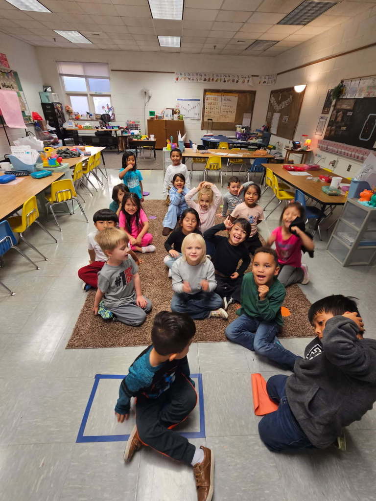 “Wide shot of the class seated on the rug for circle time; two students are in front near a taped floor square, with tables and shelves around the room.”