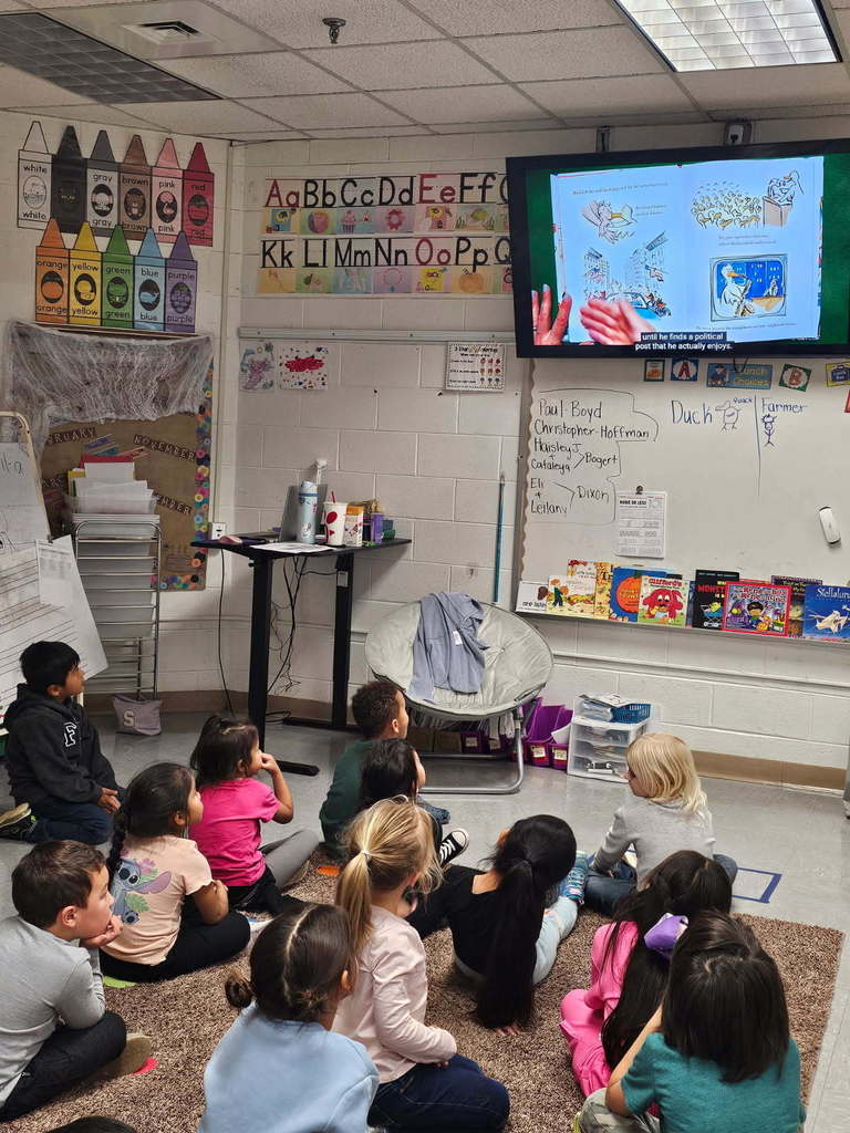 “Class gathered on the rug watching a story on a wall-mounted screen; alphabet posters and a row of picture books are visible at the front of the room.”