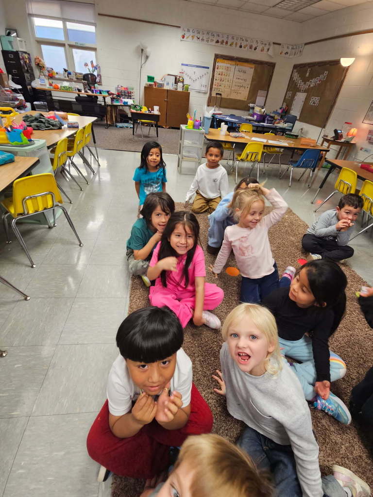 “Small group of students seated on a rug, smiling up toward the camera in an elementary classroom.”