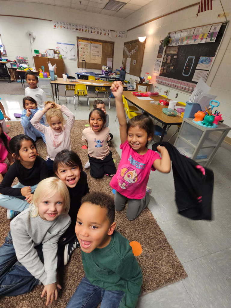 “Students on the rug cheering and smiling with hands raised, showing excitement during a class activity.”