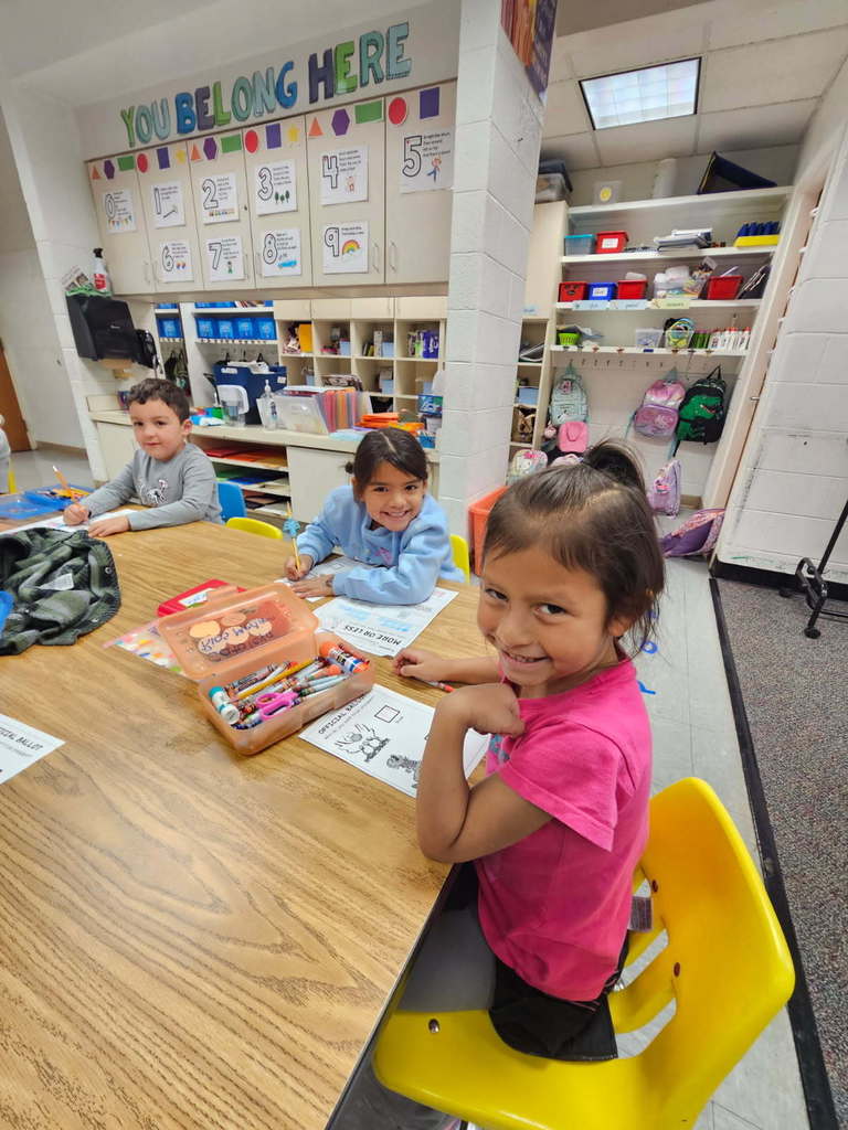 “Three students seated at a table, smiling and completing worksheets; a ‘You Belong Here’ sign and storage cabinets are behind them.”
