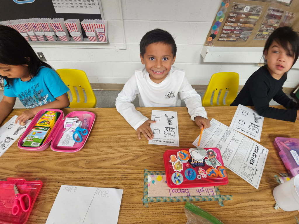 Students at a table completing worksheets with pencil boxes and supplies; the center student smiles toward the camera.”