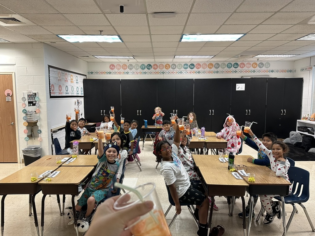 “Students sitting at desks in a classroom wearing pajamas and raising cups in celebration, smiling toward the camera.”
