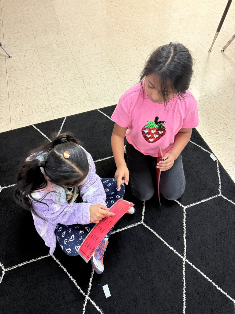 “Two students sitting on a black carpet with white lines, working together as one points to words on a red paper strip.”