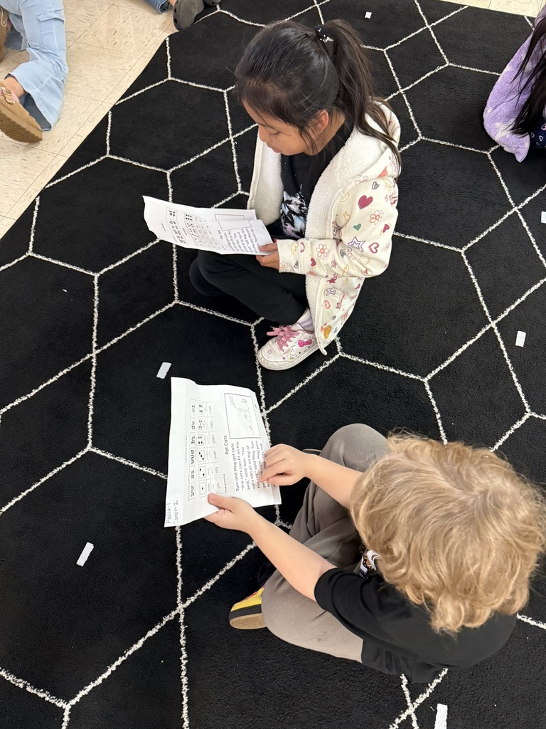 Student sitting on a black carpet with white geometric lines, reading and pointing to text on printed worksheets.”