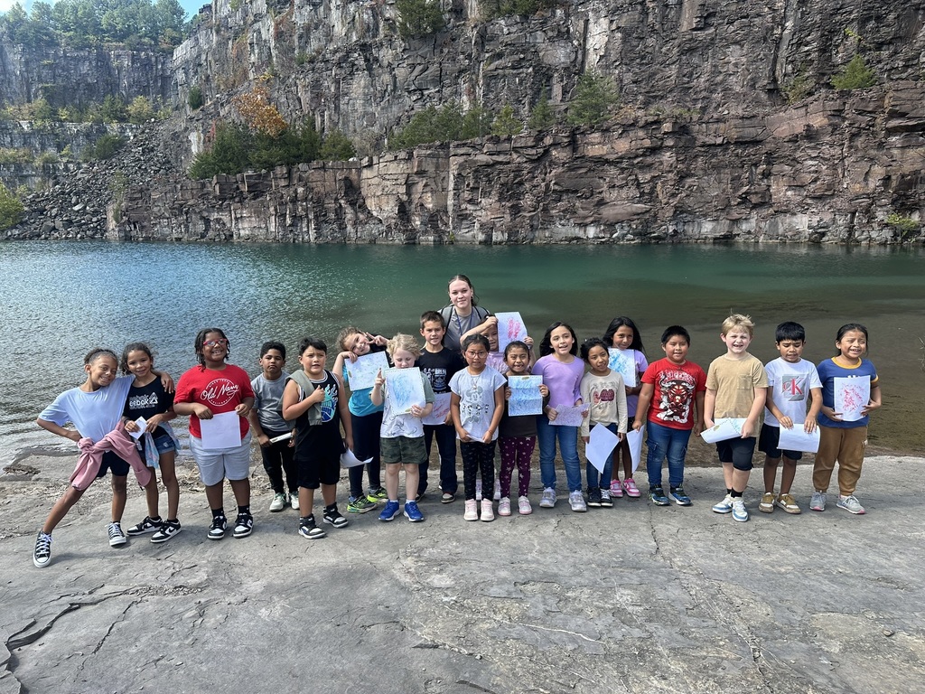 “Group of children standing with a teacher near the water at a rock quarry, holding up papers from their fossil rubbing activity.”
