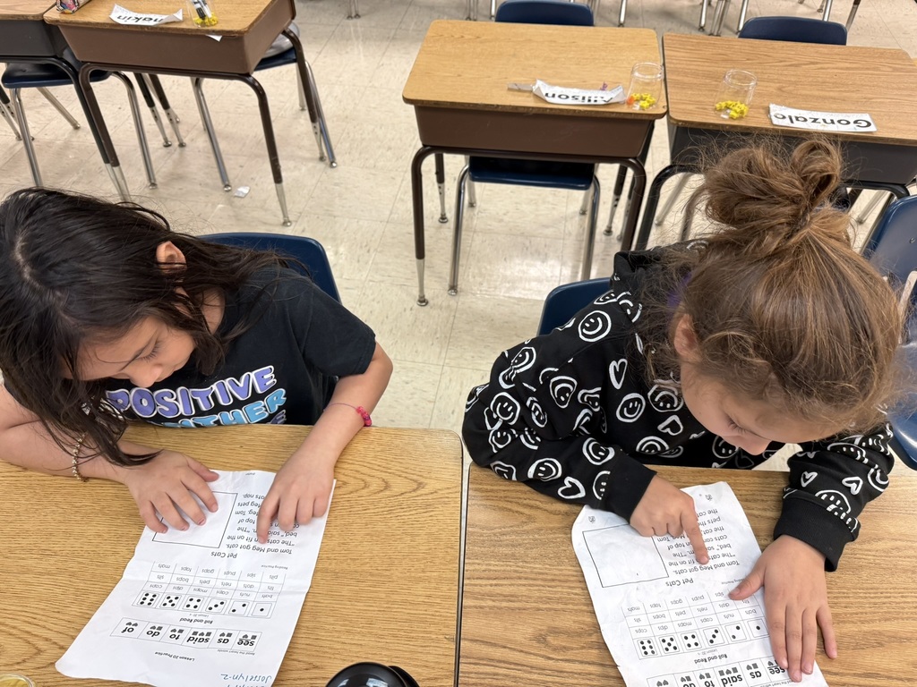 “Two students at a classroom table reading and tracking words with their fingers on a printed literacy worksheet.”