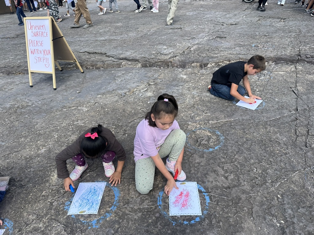 “Students crouching on stone ground making rubbings with crayons over fossil imprints; a sign nearby reads ‘Uneven Surface Please Watch Your Step.’”