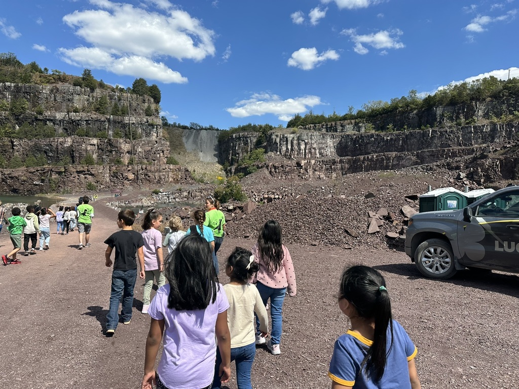 “Students walking in a line through a large rock quarry under a blue sky with scattered clouds, accompanied by adults in bright green shirts.
