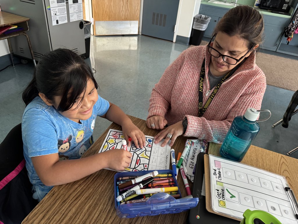 “Student and teacher sitting together at a table, smiling while working on a colorful worksheet using markers and crayons.