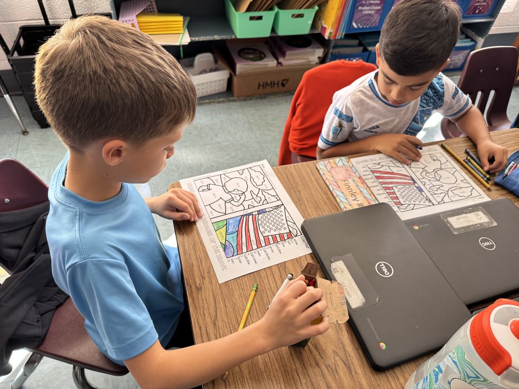 “Two students sitting side by side at a table, coloring U.S. map worksheets with bright crayons and pencils; Chromebooks and water bottles nearby.”