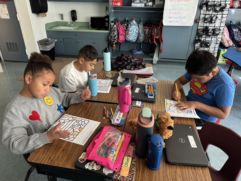 “Three students sitting at a table coloring math activity sheets with pencils and crayons; school supplies and laptops on the table.”