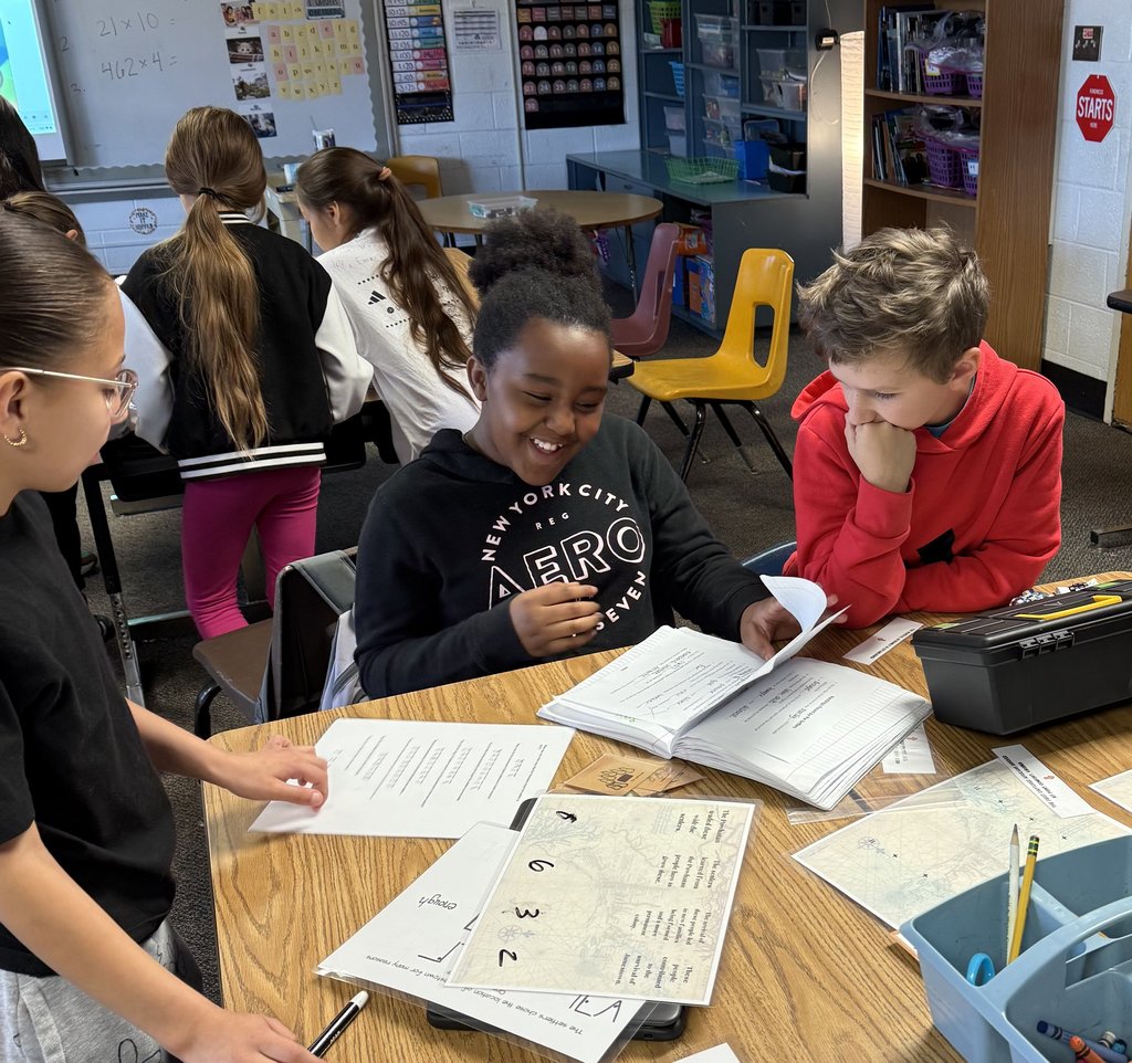 “Three students working together and smiling as they look through notebooks and worksheets during a classroom problem-solving activity.”