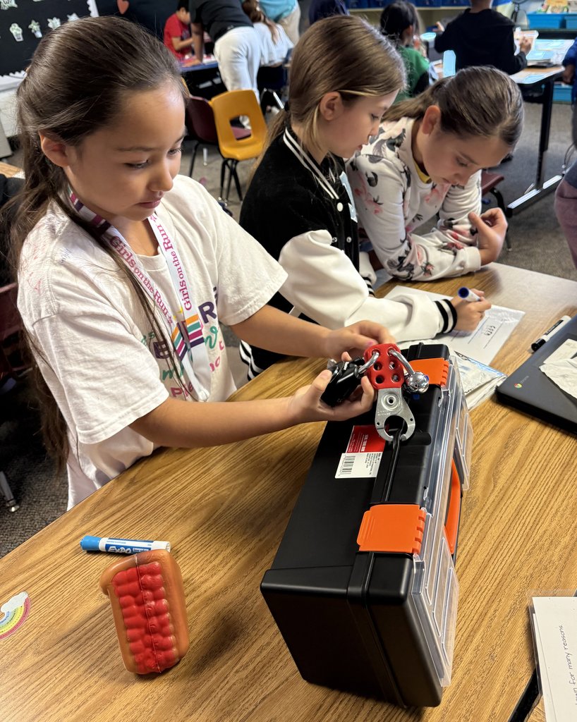 “Student examining a red lock mechanism attached to a toolbox while classmates beside her write clues on paper.”