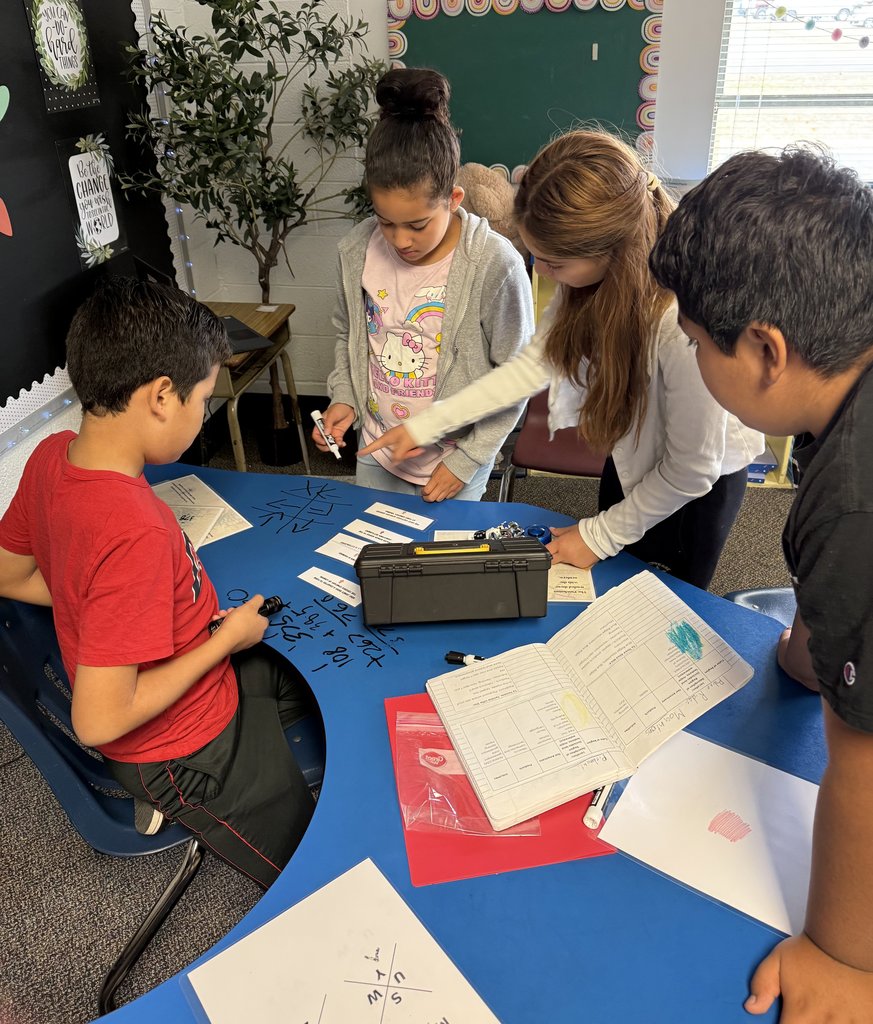 “Four students collaborating at a blue table, solving math or word puzzles with notebooks, markers, and a lockbox nearby.”