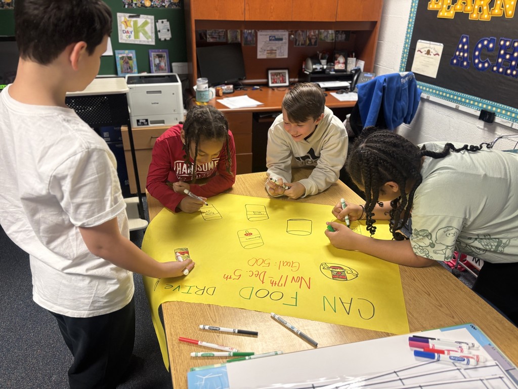 4 students work around a table in a classroom on a yellow sheet of bulletin board paper- Reads CAN FOOD DRIVE Nov 17-Dec 5 GOAL 500
