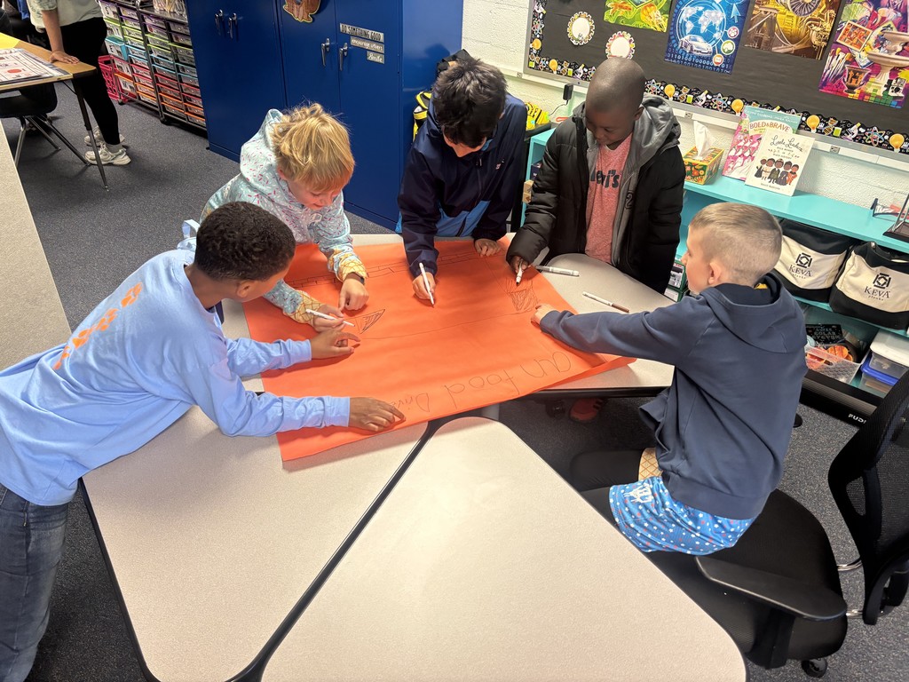 A group of 5 students work around a table with a large sheet of orang bulletin board paper.