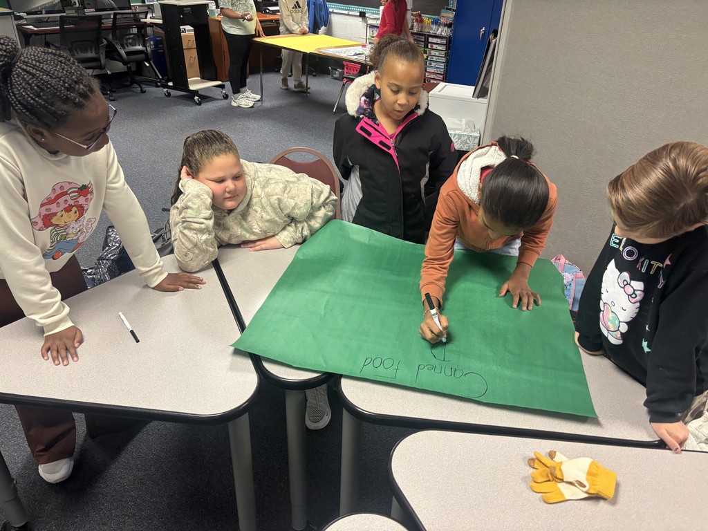 A group of 5 students work around a table with a large sheet of green bulletin board paper- CANNED FOOD