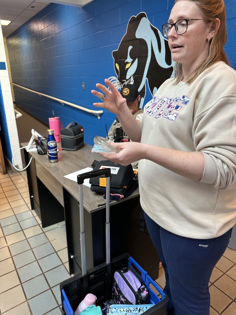 A woman wearing glasses and a light sweatshirt stands beside a rolling supply cart, holding medical equipment and speaking as if giving instructions. Behind her is a blue wall with a large panther mascot graphic and a seated officer.