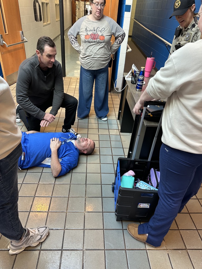 A man lies on the floor in a school hallway, appearing to participate in a medical or safety training scenario. Several adults stand or kneel around him observing, including a woman in a “thankful, grateful, blessed” pumpkin-themed shirt and a uniformed officer. A rolling cart with medical supplies and bags is nearby.