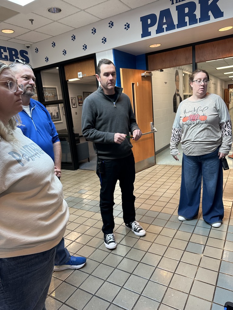A group of adults stands in a school hallway near the entrance to an office area labeled “The Park.” They appear to be listening to someone speaking, likely as part of a training or demonstration. The tiled floor and open wooden doors frame the group.