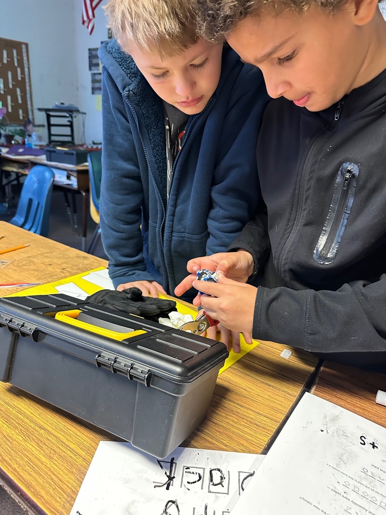 Students receive guidance from an adult pointing at clue cards on the table. A toolbox, laminated worksheets, and lockboxes are part of the activity.