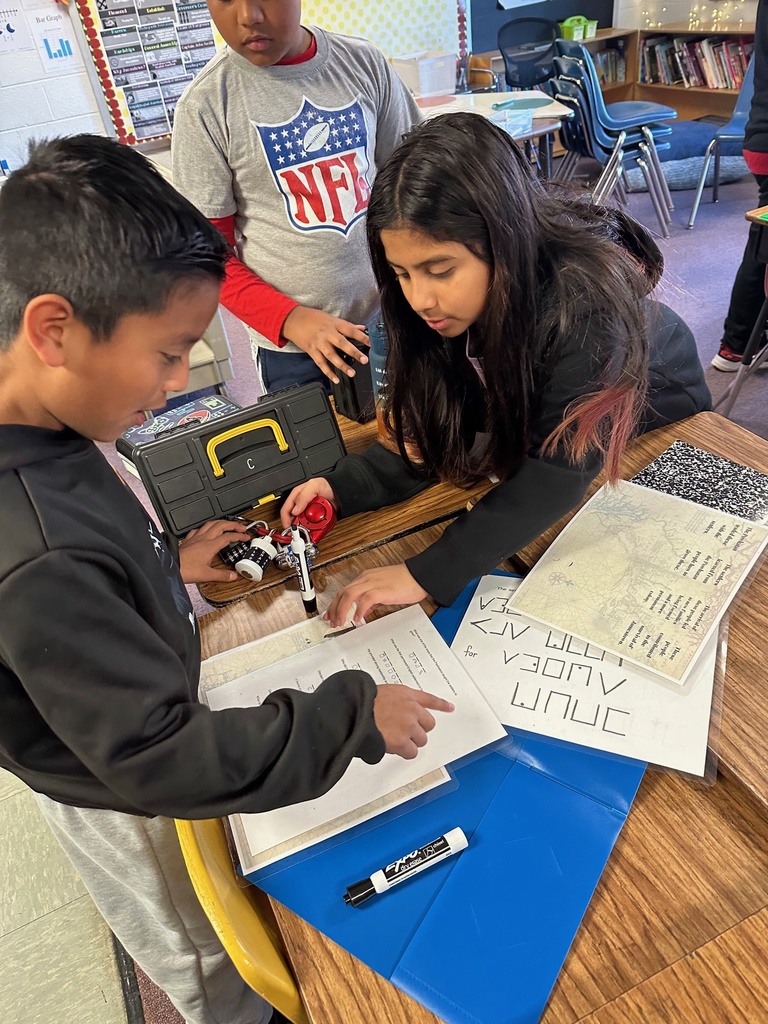 A student in a striped sports jersey holds a small metal lockbox while another student works on puzzle sheets and a folder on the table.