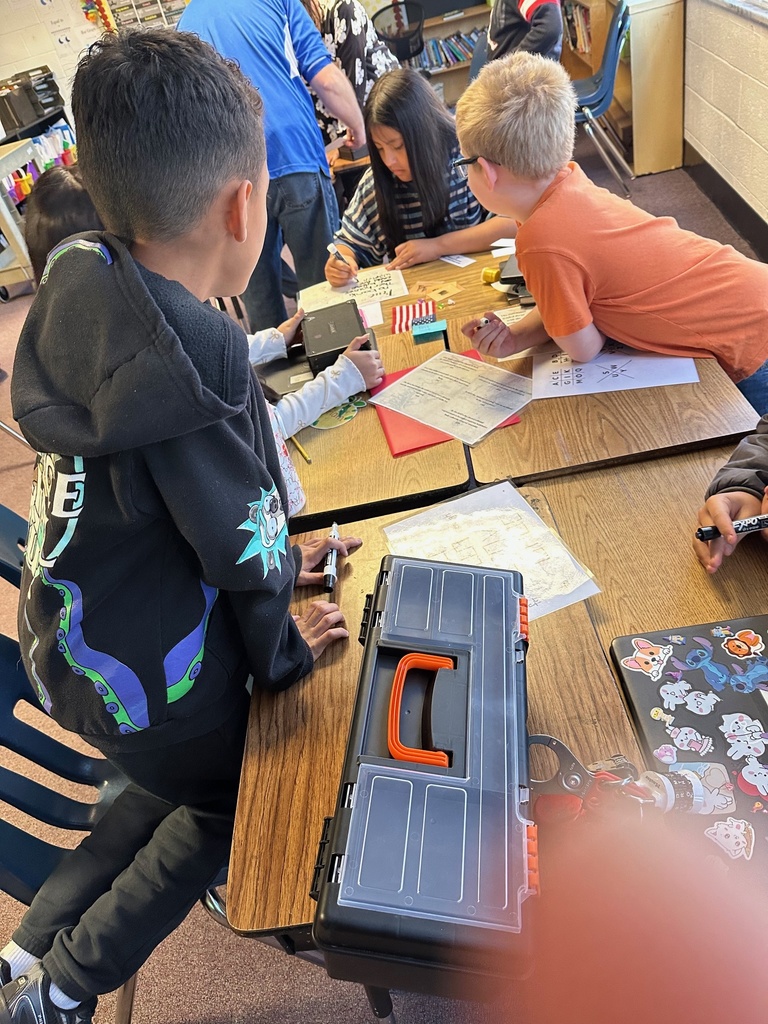 Two students lean closely over a black toolbox, examining a small object or clue. Papers with coded symbols are on the table next to them.