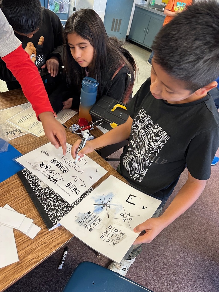Students receive guidance from an adult pointing at clue cards on the table. A toolbox, laminated worksheets, and lockboxes are part of the activity.