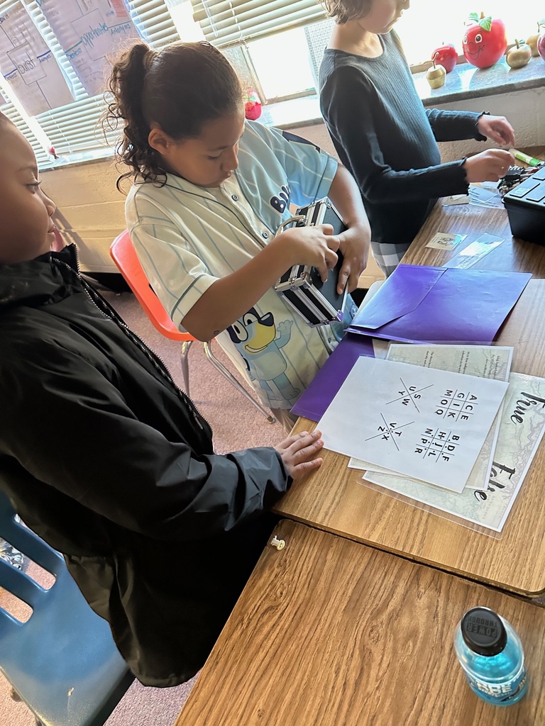 A student holds a sheet with cipher-style symbols while another student points to clues on the table. Several laminated puzzle sheets and a composition notebook are nearby as the group works together.