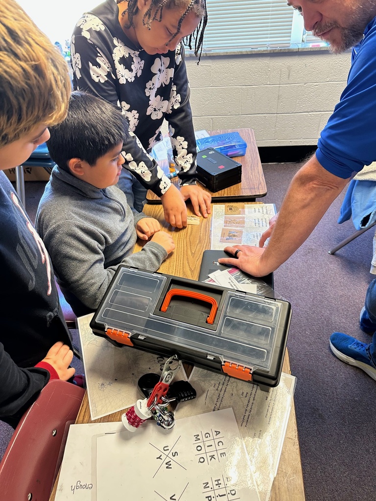 Students receive guidance from an adult pointing at clue cards on the table. A toolbox, laminated worksheets, and lockboxes are part of the activity.