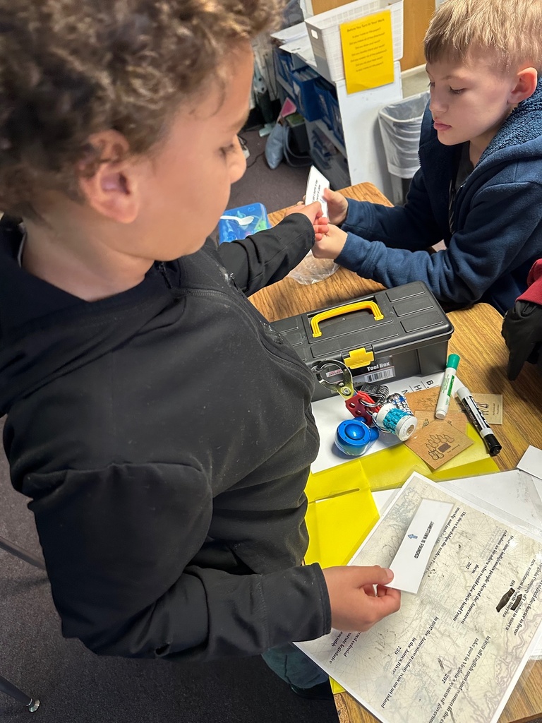 Students gather around a toolbox on a classroom table, using their hands to manipulate a lock and clues. Worksheets with numbers and images are spread out beside them.