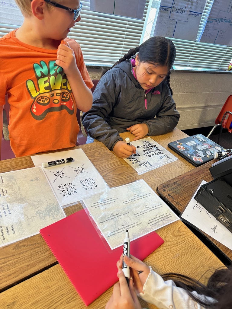 Students work at a table with laminated clue sheets, a bright pink folder, and dry-erase markers. One student writes on a sheet while others read and discuss the puzzle.