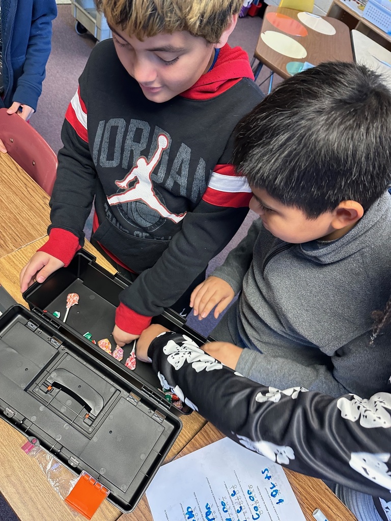 Students look inside an open toolbox filled with colorful lollipops. Hands reach in as they search through items for a puzzle or clue.