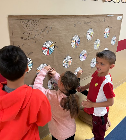 Three students stand in front of the same brown bulletin board covered in colorful pizza-slice multiplication wheels. One student is placing their circle on the board while the others watch and smile.