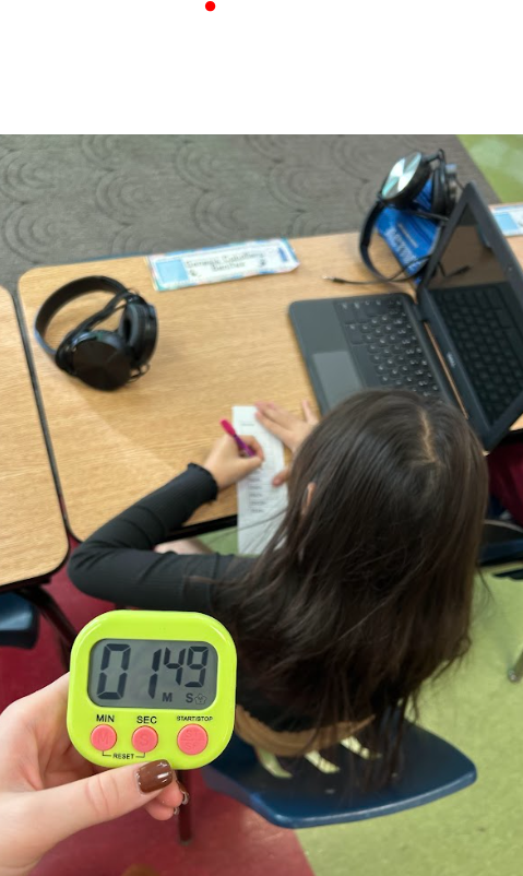 A student is seated at a desk working on a small slip of paper while a laptop and headphones sit nearby. In the foreground, a hand holds a bright green digital timer showing 01:49.