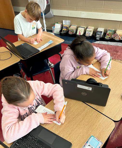 Three students sit at their desks working on small slips of paper. Laptops, headphones, and desk name tags are visible as they concentrate on the activity.