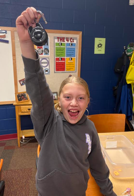 girl in a gray hoodie beams with excitement as she holds a large black combination lock triumphantly above her head.