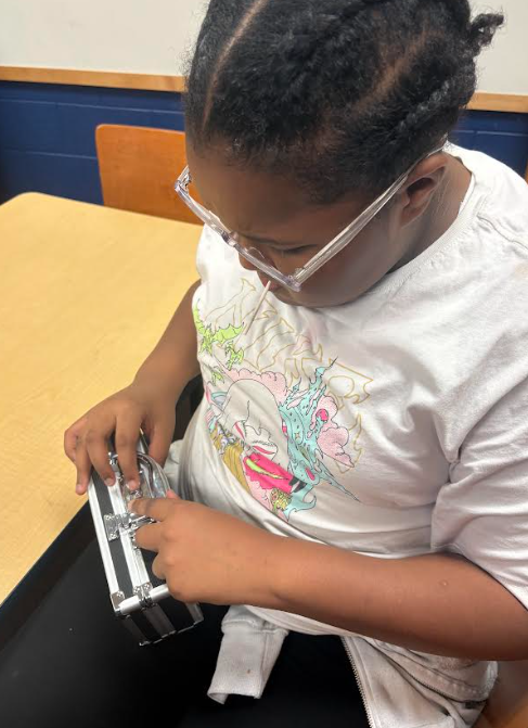 A student wearing white glasses and a white shirt focuses intently as she works to open a silver lockbox on the table.
