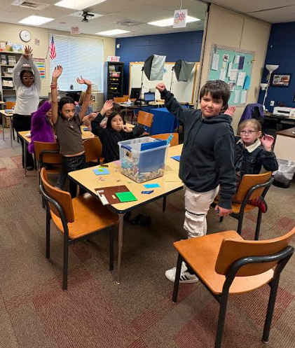 A group of students stand around a table, many with their hands raised in excitement. A large bin of puzzle materials sits on the table in front of them.