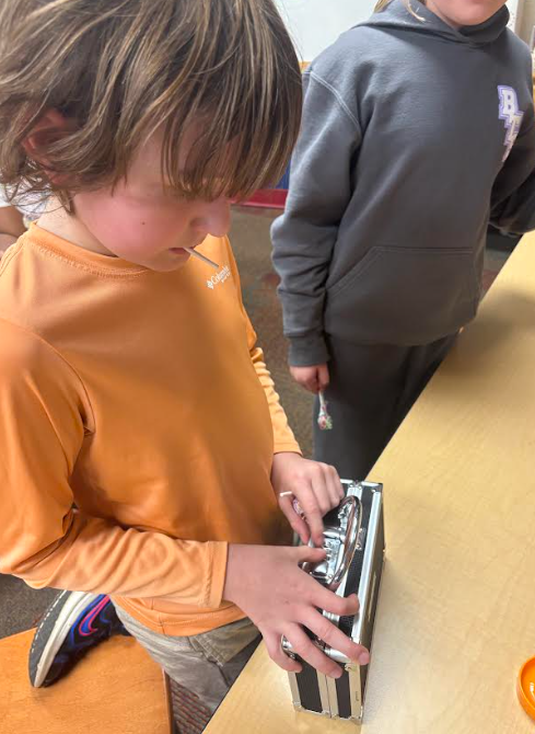 A student in an orange long-sleeved shirt concentrates as he tries to open a metal lockbox on the table. Another student stands next to him holding a lollipop.