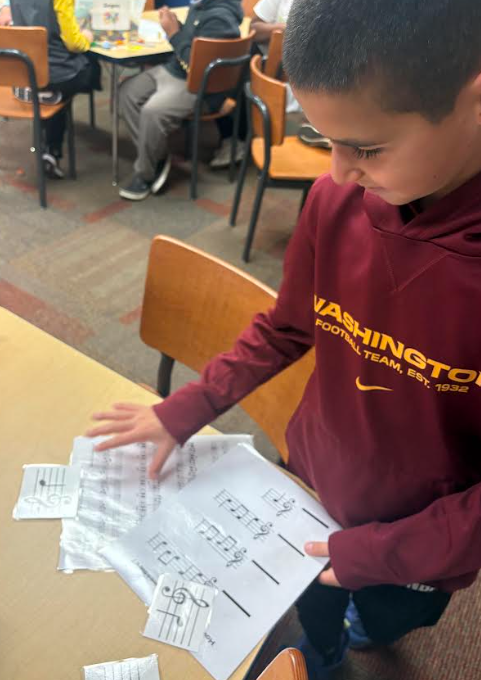 A fifth-grade boy wearing a burgundy Washington football sweatshirt stands at a table, sorting through scattered music-note puzzle pieces printed on white paper. Other students sit at tables working in the background.