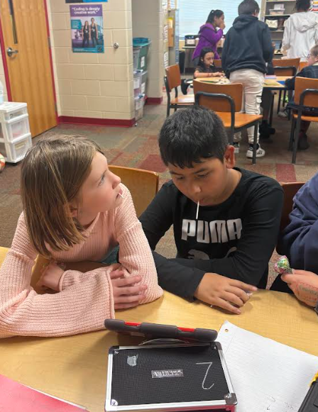 Two students sit side-by-side at a table, focused on a tablet in front of them. The girl in a light pink sweater leans in toward the boy in a black Puma shirt as they work together.