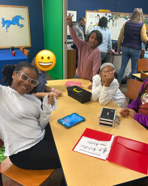A group of four students sit around a table smiling and celebrating. One student holds up a lollipop, and another smiles widely beside a tablet and a black lockbox. A yellow smiley face covers one student’s face for privacy.