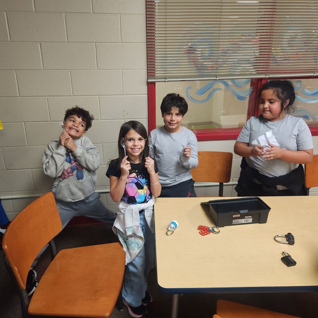 Four smiling students stand behind a table holding up metal spoons they unlocked from the Breakout boxes. Locks and clue materials are on the table in front of them.