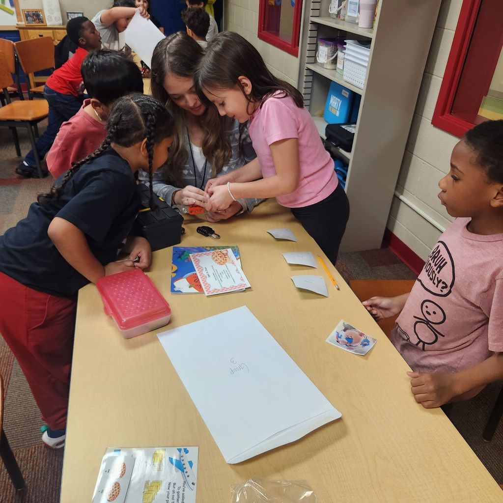 A teacher kneels beside three students as they work on unlocking a Breakout EDU lock. The group studies clue cards and a book while another student watches from the side.